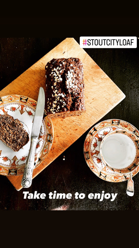 A cup and saucer of tea beside a side plate holding a slice of Stout City Loaf, with the whole loaf on a wooden chopping board and the words “Take time to enjoy” beneath